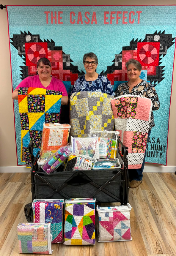 Three women stand in front of a large quilt reading 'The CASA Effect,' holding colorful donated quilts, with a wagon full of quilts and packaged quilt sets in front of them.