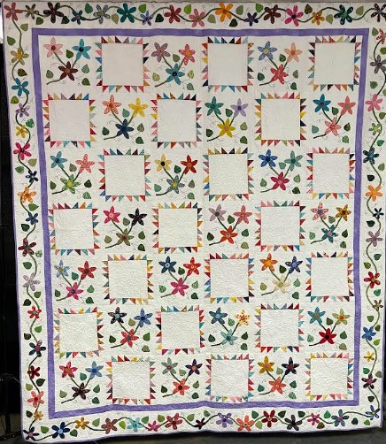 The guild's 2027 raffle quilt: white blocks framed by colorful sawtooth points, scattered appliqué flowers with green vines, a lavender inner border, and a flowering vine outer border.